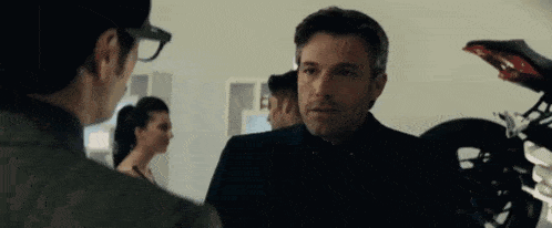 a man in a suit and glasses is talking to another man in front of a motorcycle in a room .