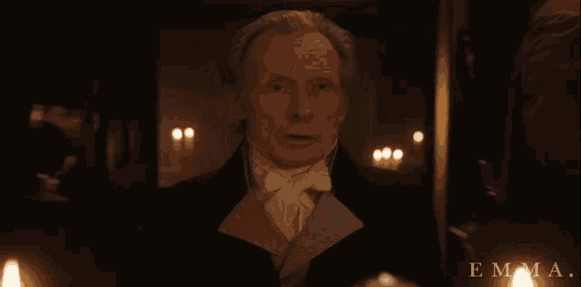 a man in a suit stands in a dark room with candles and the word emma on the bottom
