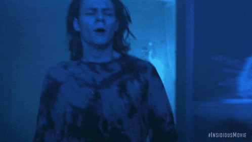 a man in a tie dye shirt is standing in a dark room with #insidiousmovie written on the bottom right