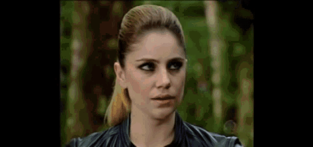 a woman in a black leather jacket is standing in the woods looking at the camera .