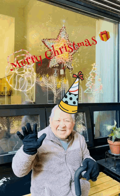 an elderly man wearing a party hat is waving in front of a window that has merry christmas written on it