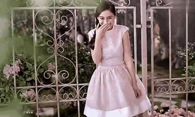 a woman in a pink dress is standing in front of a wrought iron fence covering her mouth with her hand .