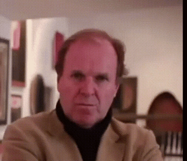a man in a tan jacket and black turtleneck looks at the camera with his arms crossed