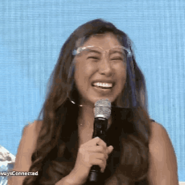 a woman wearing a face shield and glasses is laughing while holding a microphone .
