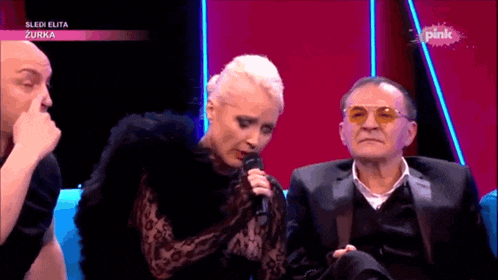 a woman is singing into a microphone while sitting next to two men on a pink screen