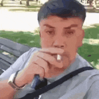 a man is sitting on a park bench smoking a cigarette .