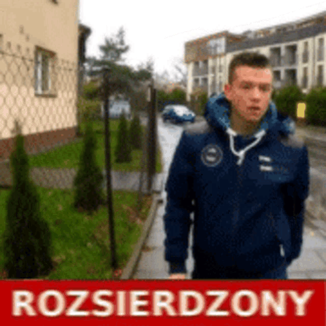 a man in a blue jacket is standing on a sidewalk in front of a sign that says rozsierdzony