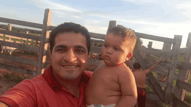 a man in a red shirt holds a baby in front of a wooden fence