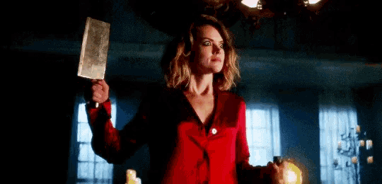 a woman in a red jacket is holding a large knife in her hand in a dark room .