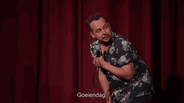 a man in a hawaiian shirt is holding a microphone on a stage and says goeiendag