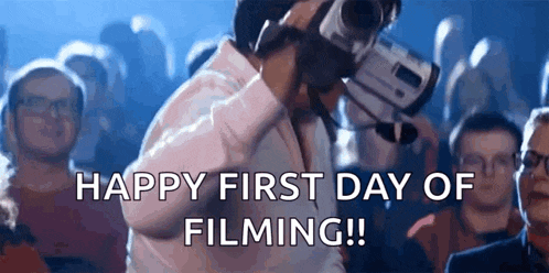 a man is holding a video camera in front of a crowd of people and says happy first day of filming !