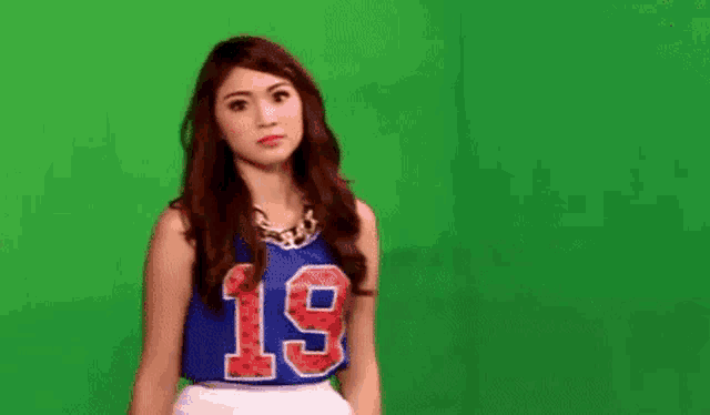 a young woman is standing in front of a green screen wearing a number 19 jersey .