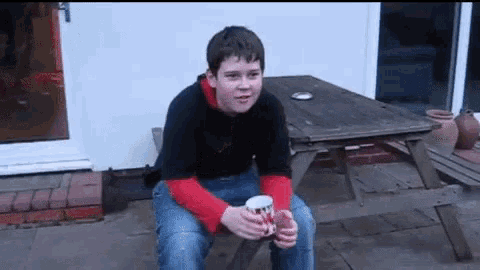 a young boy is sitting at a picnic table holding a cup of coffee