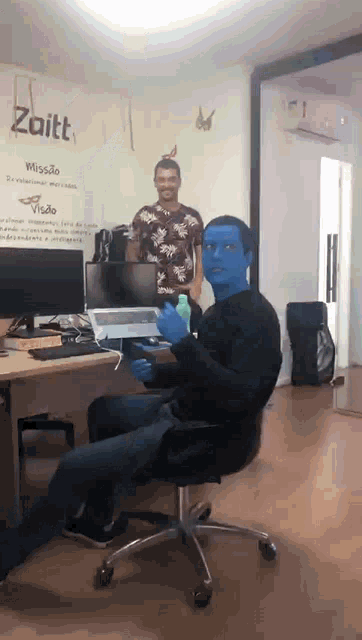 a man with blue paint on his face sits at a desk in front of a sign that says zaitt