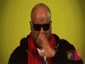 a bald man wearing sunglasses and a red scarf is blowing his nose .