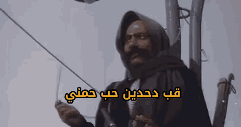 a man in a hooded jacket is standing next to a pole with arabic writing