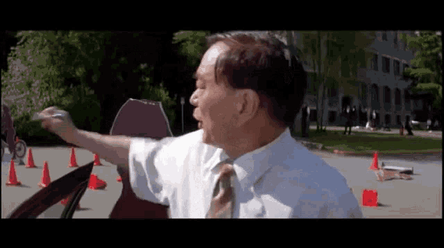 a man in a white shirt and tie is pointing at something in a parking lot .