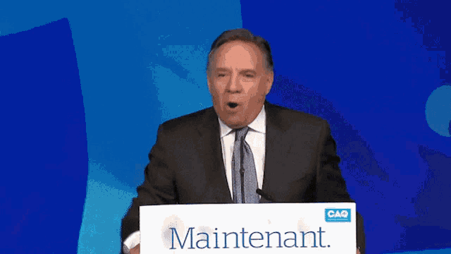 a man stands at a podium with a sign that says maintenant on it
