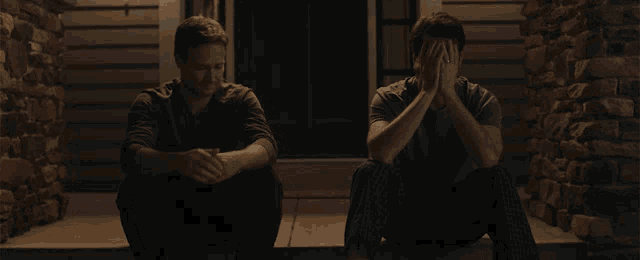 two men sit on the steps of a house with one covering his face with his hands