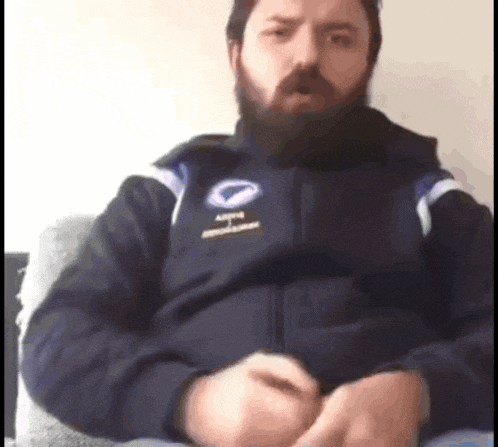 a man with a beard is sitting on a couch wearing a blue jacket with a logo for a company