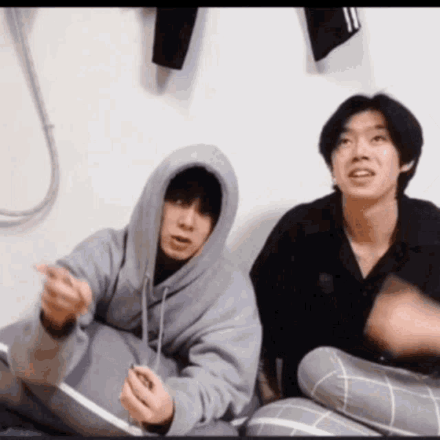 two young men are sitting next to each other on a bed . one of the men is wearing a hoodie .