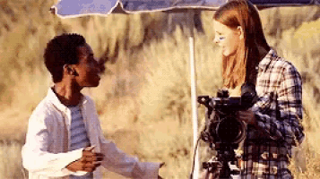 a man and a woman are standing under an umbrella talking to each other . the woman is holding a camera .