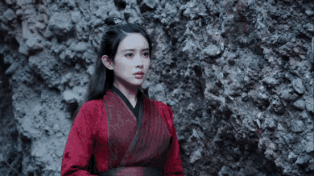 a woman in a red robe stands in front of a rock wall