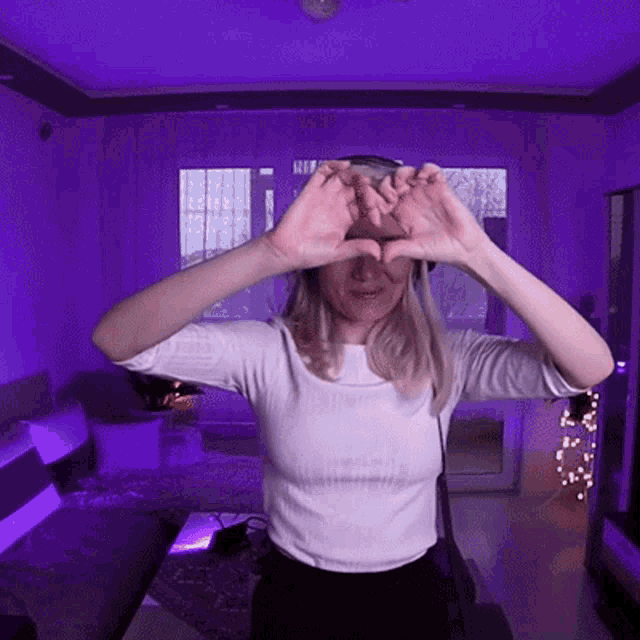 a woman is making a heart shape with her hands in a living room with purple lighting .