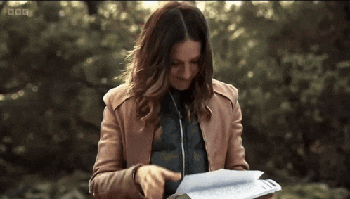 a woman in a brown jacket is looking at a piece of paper with bbc written on the bottom