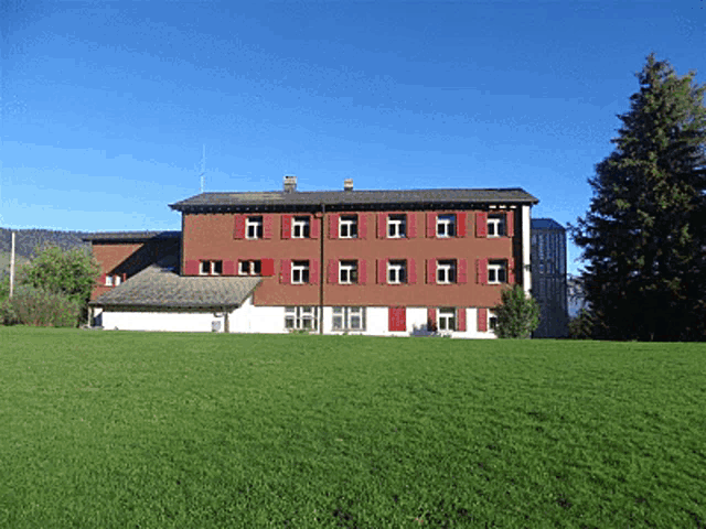 a large red building with red shutters sits in the middle of a green field