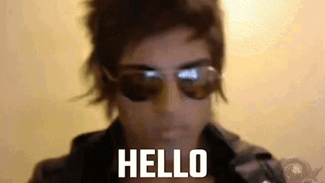 a man wearing sunglasses is standing in front of a wall and saying hello .