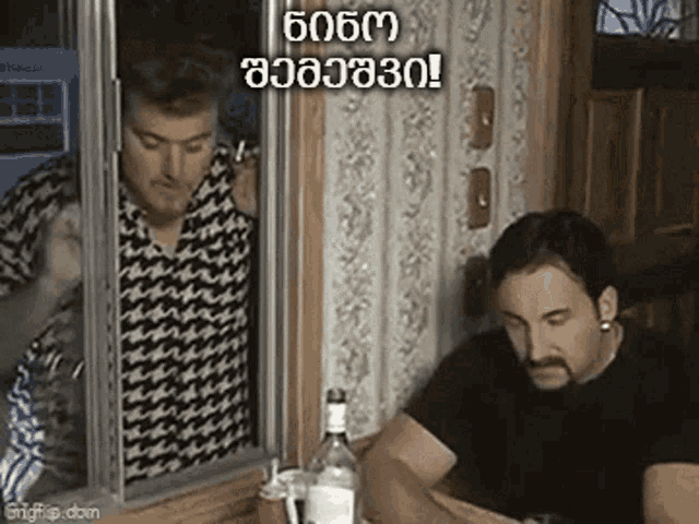 two men are sitting at a table with a bottle of alcohol and a cat looking out of a window .