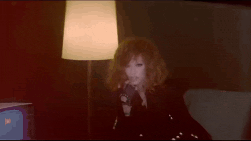 a woman with red hair is singing into a microphone in front of a lamp in a dark room .