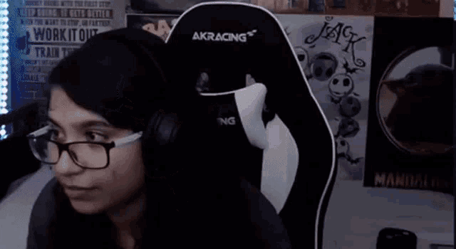 a woman wearing glasses and headphones sits in an akracing chair