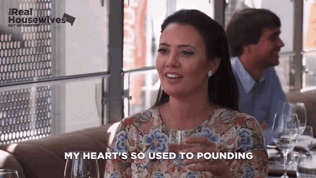 a woman is sitting at a table holding a glass of water and saying my heart 's so used to pounding