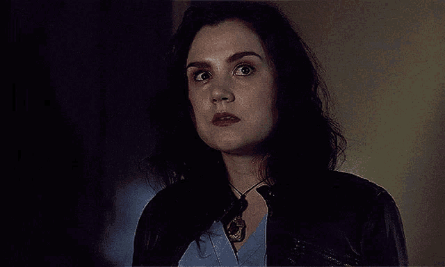 a woman wearing a blue shirt and a black jacket looks at the camera with a surprised look on her face