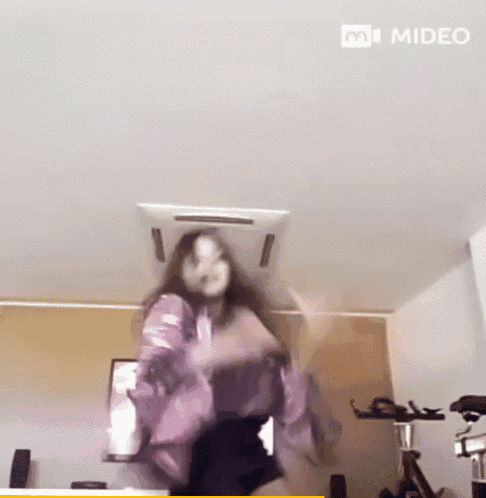 a woman in a purple shirt is jumping in the air in front of an exercise bike in a room with the words m video visible