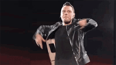 a man in a leather jacket is singing into a microphone while pointing at the camera .