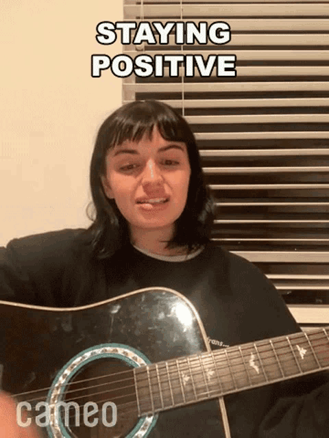 a woman is playing a guitar in front of a window with the words staying positive written above her .