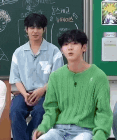 a man in a green sweater is sitting next to another man in a blue shirt in front of a blackboard .