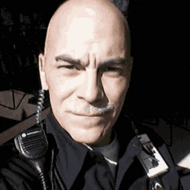 a bald man with a mustache is wearing a police uniform and a walkie talkie around his neck