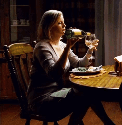 a woman sits at a table drinking from a bottle and holding a glass of wine