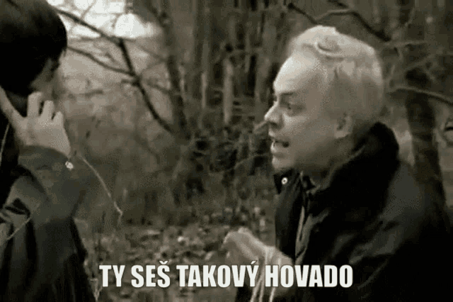 a man is talking on a cell phone in a forest while another man looks on .