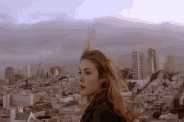 a woman is standing in front of a city skyline with her hair blowing in the wind .
