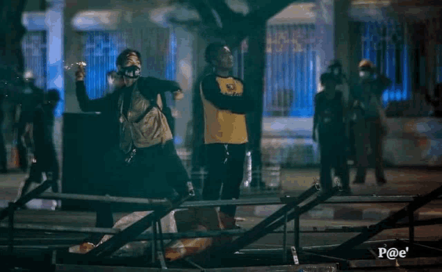 a man wearing a mask throws a bottle over a fence with the letters p @ e on the bottom right