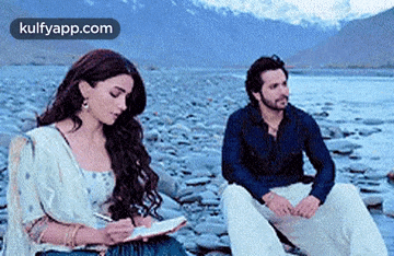 a man and a woman are sitting next to each other on a rocky beach . the woman is writing in a notebook .