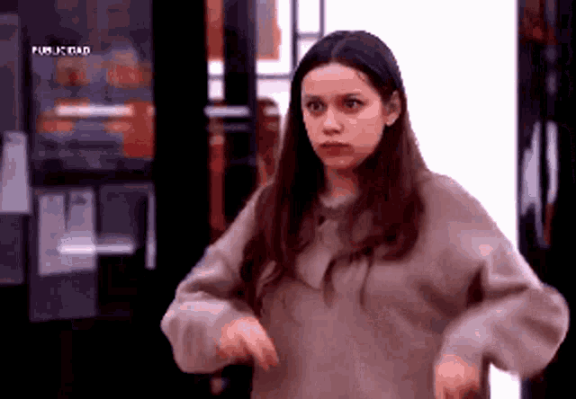 a woman in a brown sweater is standing in front of a door and making a funny face .