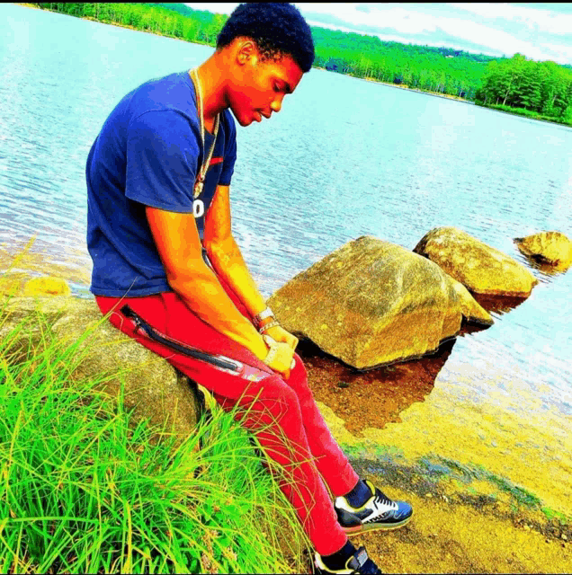 a young man sits on a rock near a lake wearing a blue shirt with the letter d on it