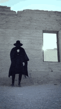 a man in a black coat and hat is standing in front of a wall