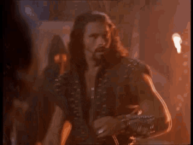 a man with long hair and a beard is standing in a dark room with a torch in the background .
